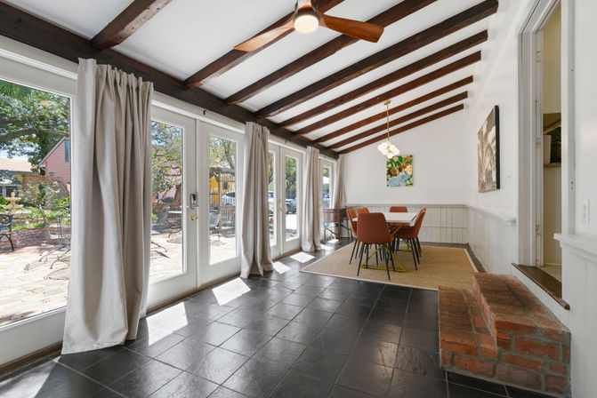 Cozy sunlit dining sunroom with exposed dark wood beams and ceiling fan, floor-to-ceiling French doors with light curtains opening to a brick patio, black tile floor, area rug and rust-colored dining chairs.