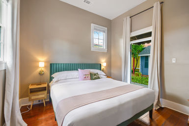 Cozy guest bedroom with emerald upholstered headboard, crisp white linens, wooden nightstand and wall sconces, warm hardwood floors and open curtains revealing a leafy garden view.