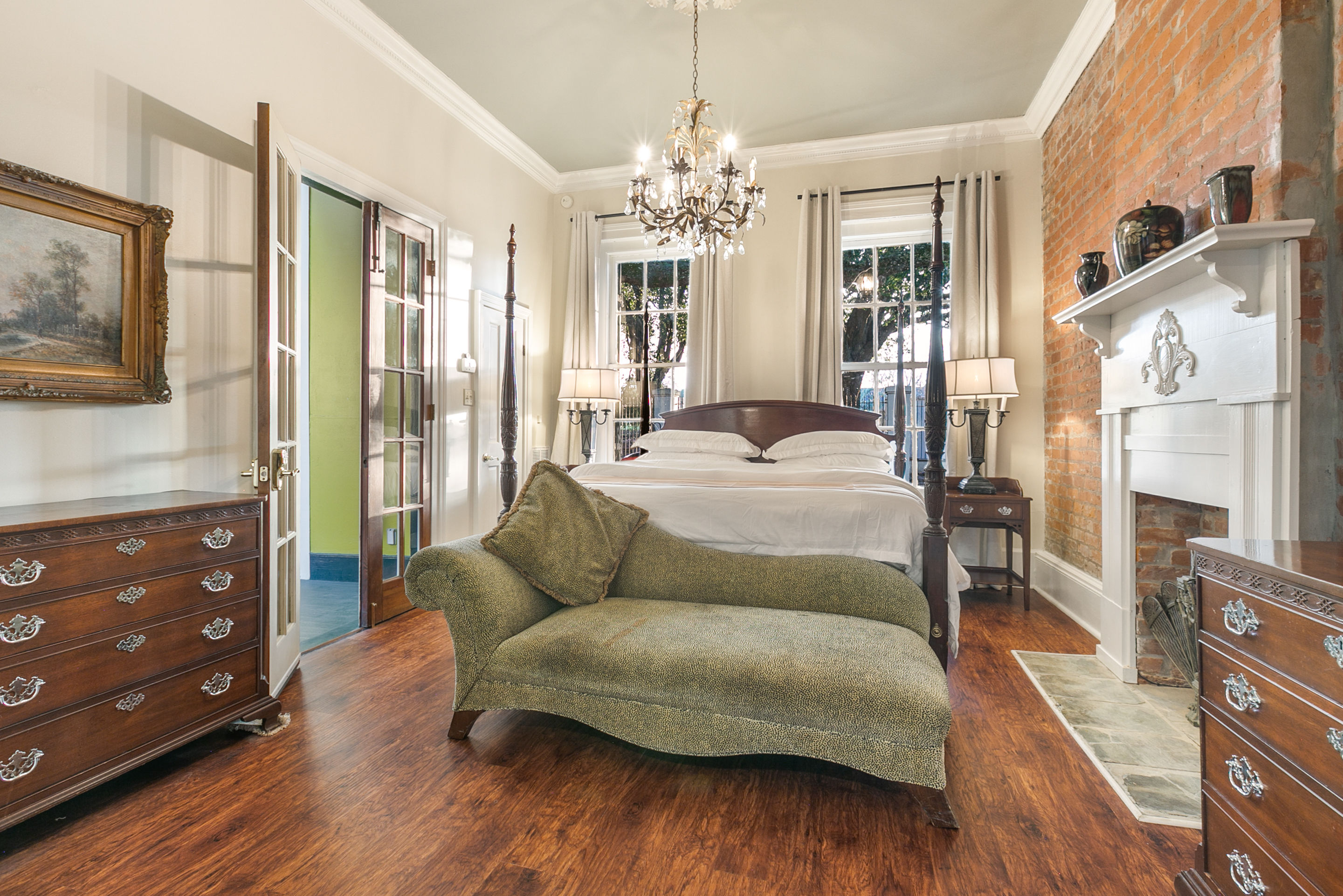 Sunlit Victorian-style bedroom with four-poster bed, green chaise lounge, crystal chandelier, exposed brick fireplace, hardwood floors, French doors and antique dressers.