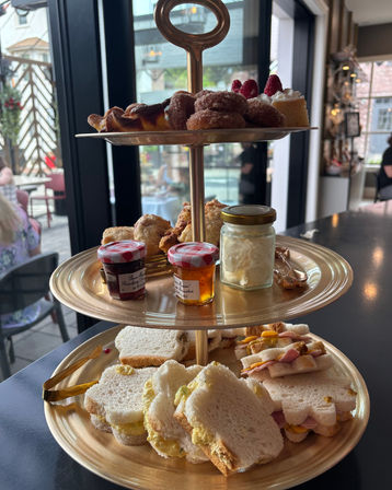 Three-tiered gold afternoon tea stand on a café table by a window with finger sandwiches, scones, jam jars, clotted cream and assorted pastries