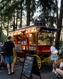 Vintage red-and-yellow popcorn wagon at a dusk park event, lit carriage serving snacks beneath tall evergreen trees with a chalkboard menu and people enjoying popcorn and cotton candy