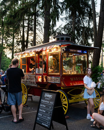Vintage red-and-yellow popcorn wagon at a dusk park event, lit carriage serving snacks beneath tall evergreen trees with a chalkboard menu and people enjoying popcorn and cotton candy
