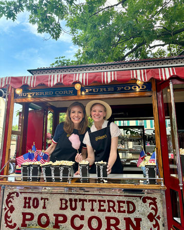 Two smiling vendors behind a vintage red-and-white striped popcorn cart serving hot buttered popcorn and a pink cotton candy, decorated with small American flags at an outdoor summer street fair.