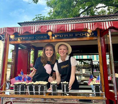 Two smiling vendors behind a vintage red-and-white striped popcorn cart serving hot buttered popcorn and a pink cotton candy, decorated with small American flags at an outdoor summer street fair.
