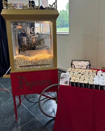 Vintage red-and-gold popcorn cart with glass kettle and overflowing popped corn next to a red-clothed snack table lined with individual popcorn cups and lids in an indoor event space.