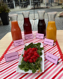 Outdoor patio table with four glass carafes labeled grapefruit, cranberry, pomegranate and orange juice on a pink-striped runner, plus a white platter of raspberries and fresh mint.