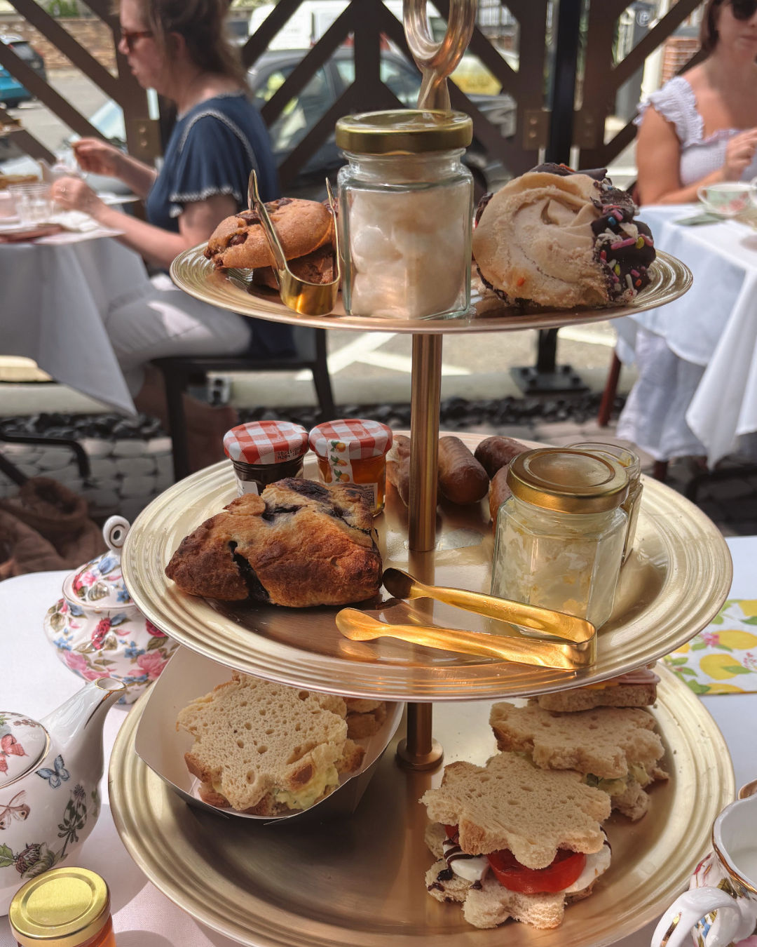 Three-tiered gold afternoon tea stand on an outdoor cafe patio with finger sandwiches, scones, jam jars and pastries, floral teapot and blurred diners in the background.