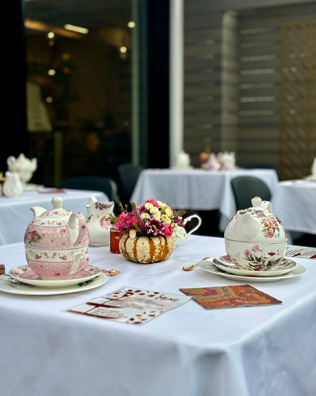 Charming outdoor afternoon tea on a white-tablecloth patio table with vintage floral teapots and teacups, a mini pumpkin vase of pink and white flowers, menus and gold spoons.