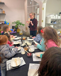 Instructor leading a kids' baking workshop in a cozy kitchen studio, children gathered around a table with bowls of dry ingredients, wooden spoons, and recipe sheets.