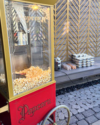 Red vintage popcorn cart filled with buttered popcorn on a cobblestone patio beside a bench with stacked foil trays, dipping cups and sauce bottles against a decorative wooden lattice wall.