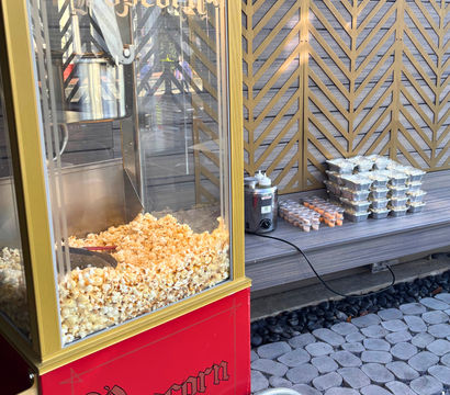 Red vintage popcorn cart filled with buttered popcorn on a cobblestone patio beside a bench with stacked foil trays, dipping cups and sauce bottles against a decorative wooden lattice wall.