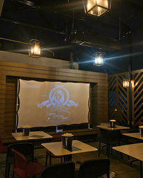 Cozy outdoor patio movie setup at night with a projected vintage film logo on a screen, warm lantern-style hanging lights, small tables and chairs, and popcorn buckets on each table.