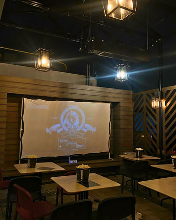 Cozy outdoor patio movie setup at night with a projected vintage film logo on a screen, warm lantern-style hanging lights, small tables and chairs, and popcorn buckets on each table.