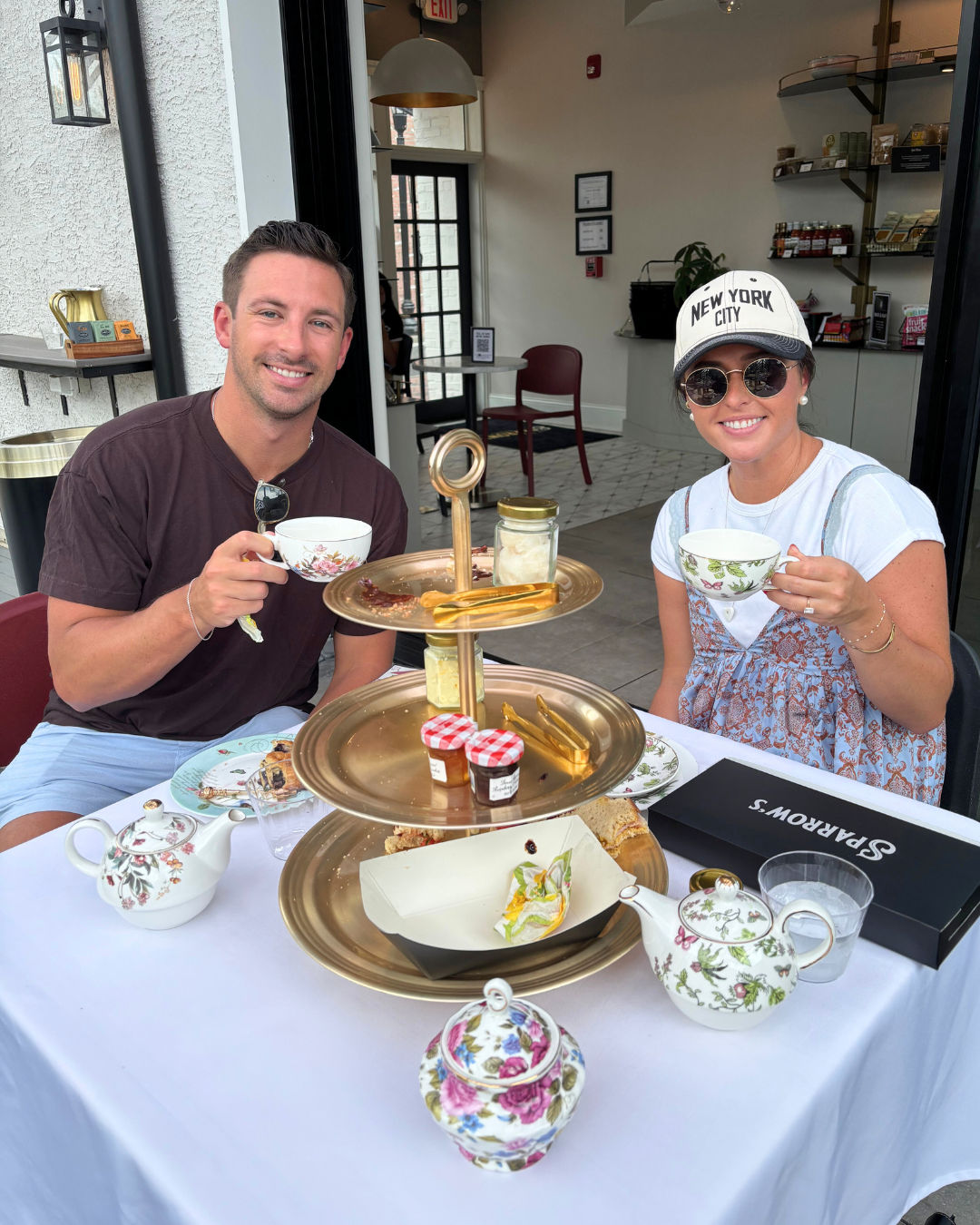 Two people enjoying outdoor afternoon high tea at a sidewalk café, smiling with floral teacups and a three-tiered tray of scones, jams and pastries.