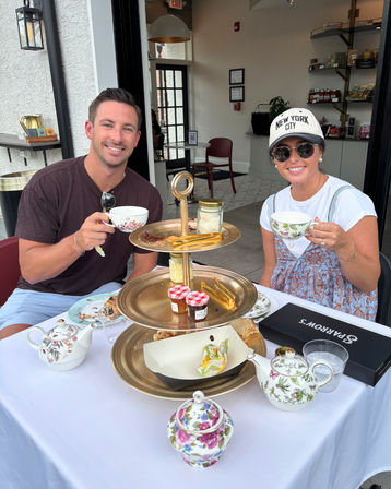 Two people enjoying outdoor afternoon high tea at a sidewalk café, smiling with floral teacups and a three-tiered tray of scones, jams and pastries.