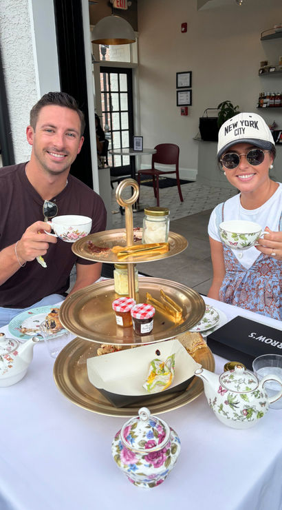 Two people enjoying outdoor afternoon high tea at a sidewalk café, smiling with floral teacups and a three-tiered tray of scones, jams and pastries.