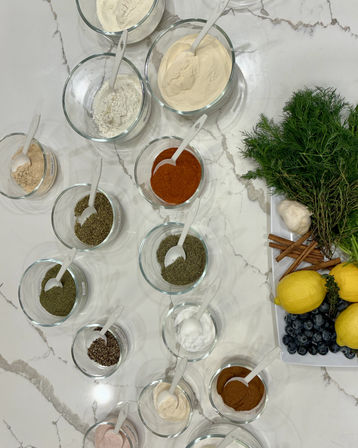 Flat-lay on a white marble kitchen countertop of glass bowls filled with assorted spices and dry powders with white spoons, next to a tray of fresh dill and herbs, garlic, cinnamon sticks, lemons and blueberries — colorful culinary ingredients.