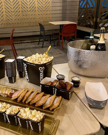 Outdoor patio snack spread with popcorn in black boxes, rows of glazed pastries and pretzel bites with dipping sauces, and wine and champagne chilling in a hammered metal ice bucket.