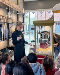 Staff member in a black apron scoops fresh popcorn from a vintage popcorn machine for a group of excited children inside a bright boutique popcorn shop with gift jars on shelves.