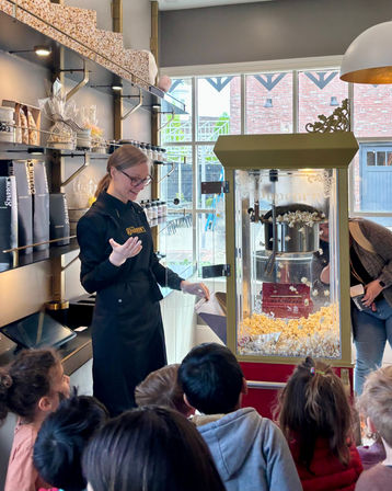 Staff member in a black apron scoops fresh popcorn from a vintage popcorn machine for a group of excited children inside a bright boutique popcorn shop with gift jars on shelves.