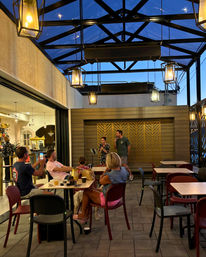 Evening outdoor patio dining under a glass-roofed pergola with hanging lanterns, live music duo on a small stage, patrons at tables (one filming) and a dog under a chair, cozy relaxed atmosphere.