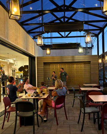 Evening outdoor patio dining under a glass-roofed pergola with hanging lanterns, live music duo on a small stage, patrons at tables (one filming) and a dog under a chair, cozy relaxed atmosphere.