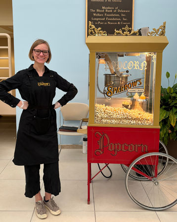 Smiling vendor in a black apron standing beside a vintage red-and-gold popcorn cart filled with freshly popped popcorn in a bright indoor space.