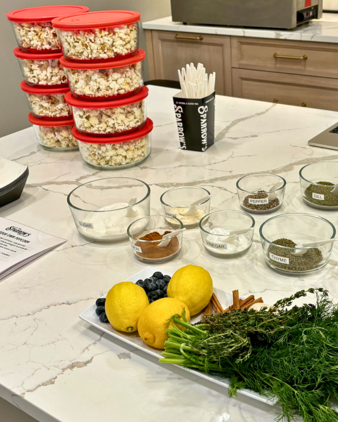 Marble kitchen island prep station with stacked clear containers of popcorn with red lids, glass bowls of labeled spices and seasonings, a cup of white spoons, and a platter of lemons, blueberries, cinnamon sticks and fresh herbs (dill, thyme).