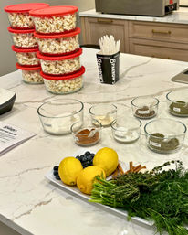 Marble kitchen island prep station with stacked clear containers of popcorn with red lids, glass bowls of labeled spices and seasonings, a cup of white spoons, and a platter of lemons, blueberries, cinnamon sticks and fresh herbs (dill, thyme).