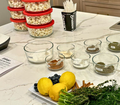 Marble kitchen island prep station with stacked clear containers of popcorn with red lids, glass bowls of labeled spices and seasonings, a cup of white spoons, and a platter of lemons, blueberries, cinnamon sticks and fresh herbs (dill, thyme).