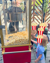 Red vintage-style popcorn machine full of freshly popped corn on an outdoor patio as a group of young children watch and reach toward the snack