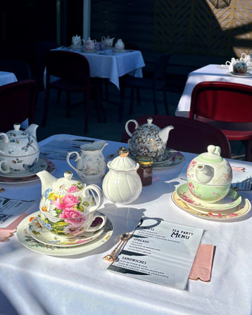 Sunlit outdoor afternoon tea on a patio with floral vintage teapots and stacked teacups, jam jar and a printed tea party menu on a white tablecloth.
