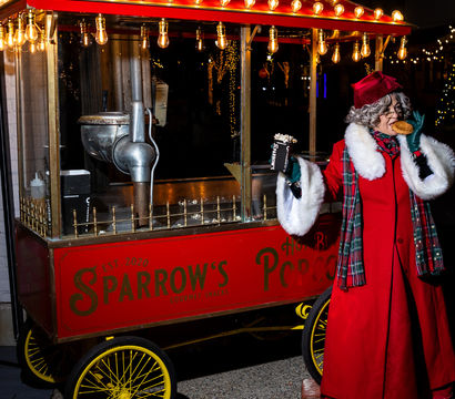 Festive nighttime street scene: person in a red coat with white-fur trim and plaid scarf and hat enjoys a snack while holding a popcorn box beside a red vintage popcorn cart lit by warm bulb lights.