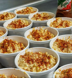 Rows of creamy Mexican street corn cups (esquites) in white takeout bowls, topped with chili-lime seasoning (Tajín) and red chili flakes — a spicy, cheesy corn snack for takeout.