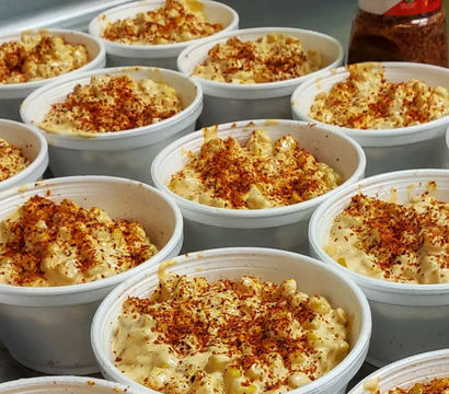 Rows of creamy Mexican street corn cups (esquites) in white takeout bowls, topped with chili-lime seasoning (Tajín) and red chili flakes — a spicy, cheesy corn snack for takeout.