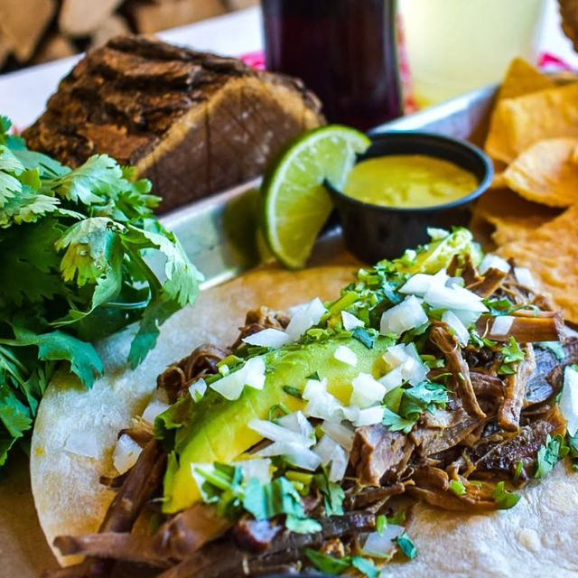 Close-up Mexican shredded beef taco on a corn tortilla topped with avocado slices, chopped onion and cilantro, served with a lime wedge, green salsa and tortilla chips.