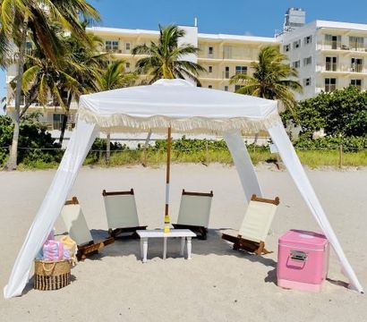 Sunny tropical beach scene with a white fringed canopy shading four low wooden lounge chairs around a small table, pink cooler and towel basket on sand, palm trees and beachfront buildings behind.