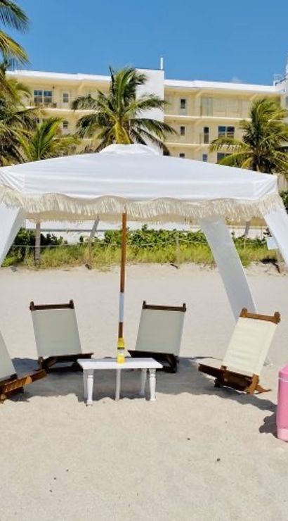 Sunny tropical beach scene with a white fringed canopy shading four low wooden lounge chairs around a small table, pink cooler and towel basket on sand, palm trees and beachfront buildings behind.
