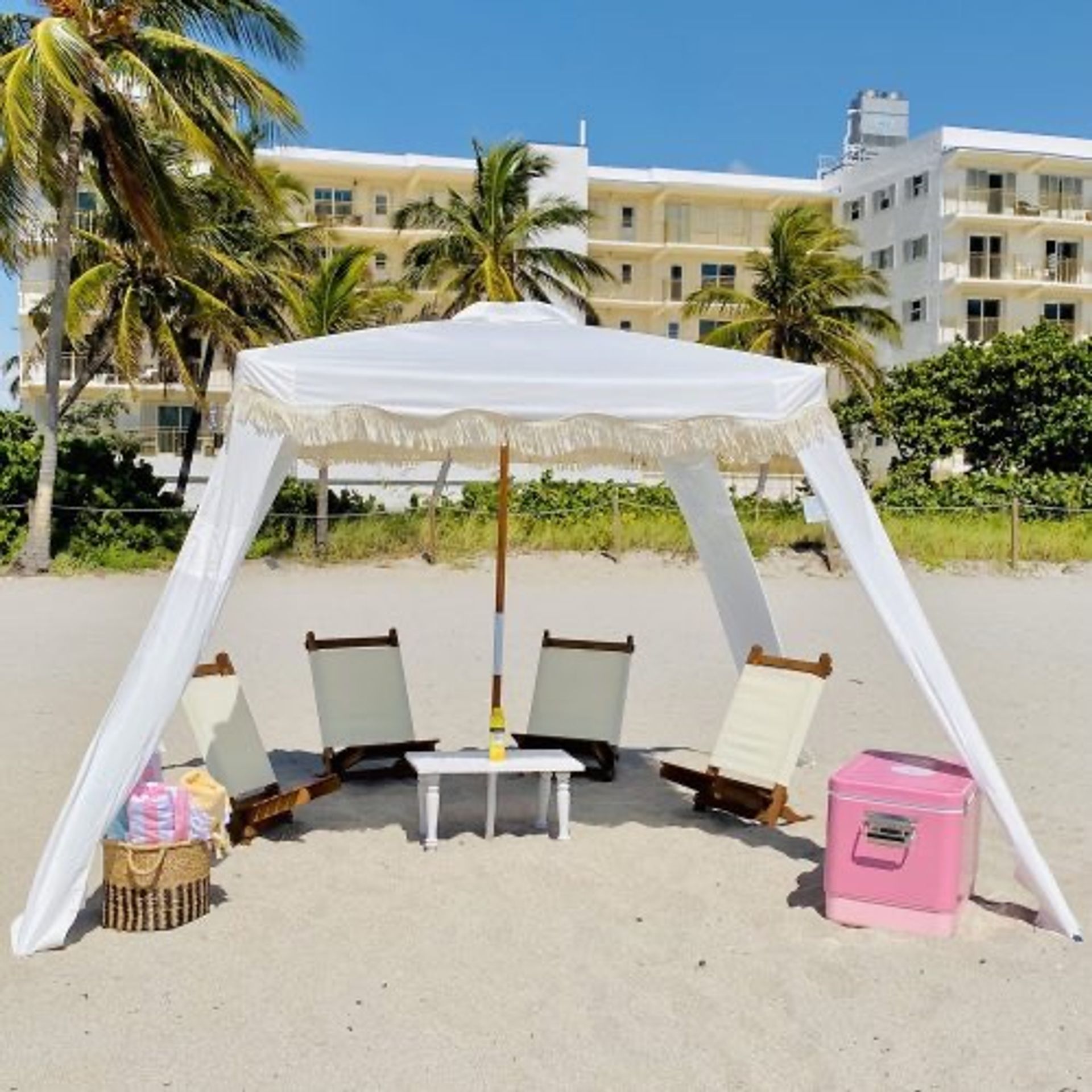 Sunny tropical beach scene with a white fringed canopy shading four low wooden lounge chairs around a small table, pink cooler and towel basket on sand, palm trees and beachfront buildings behind.