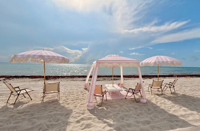 Pink-and-white striped beach umbrellas with fringe and folding chairs arranged on white sand facing a sparkling turquoise ocean under a bright blue sky — relaxed seaside scene.