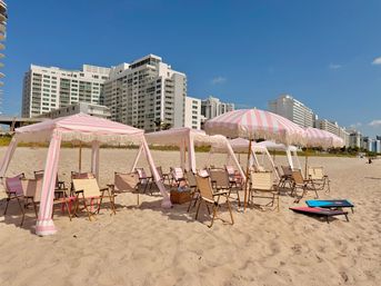 Sunny beachfront with pink-and-white striped cabanas and parasols, wooden lounge chairs on sandy shore and white high-rise condos lining the coast under a clear blue sky