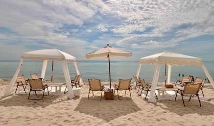 Sun-soaked beach scene with three white fringe cabanas and a central umbrella, canvas beach chairs on soft sand facing a calm turquoise ocean under a dramatic partly cloudy sky.