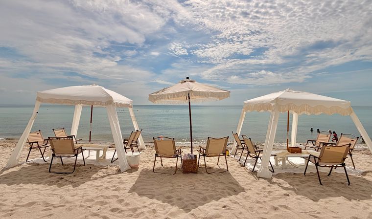 Sun-soaked beach scene with three white fringe cabanas and a central umbrella, canvas beach chairs on soft sand facing a calm turquoise ocean under a dramatic partly cloudy sky.