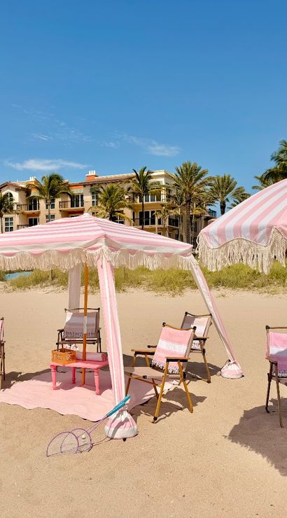 Coastal beach scene with pink-and-white striped umbrellas, a matching cabana and beach chairs on golden sand, palm trees and resort buildings under a bright blue sky.