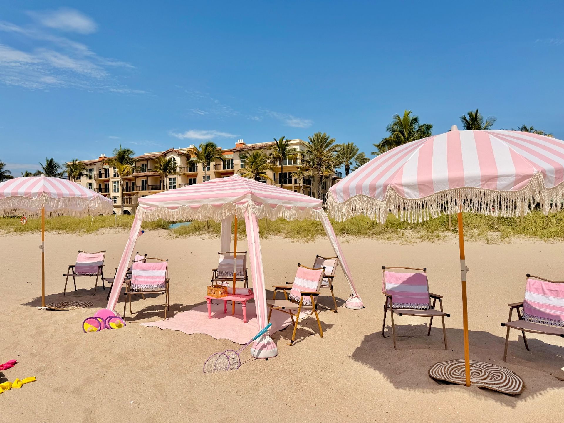Coastal beach scene with pink-and-white striped umbrellas, a matching cabana and beach chairs on golden sand, palm trees and resort buildings under a bright blue sky.