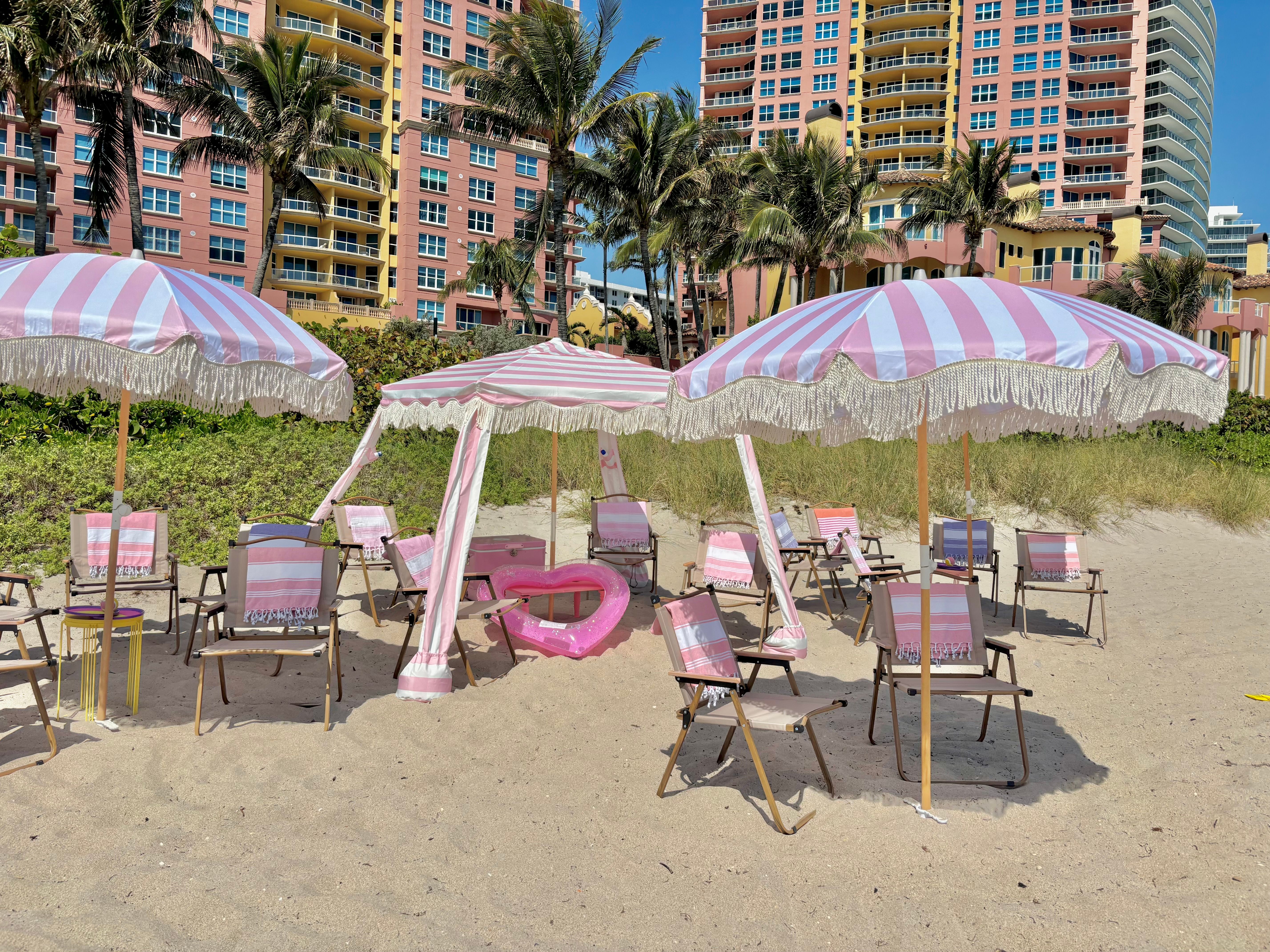 Sunny sandy beach with pink-and-white striped fringe umbrellas and matching chairs, a pink heart float, palm trees and pastel coastal high-rise buildings in the background.
