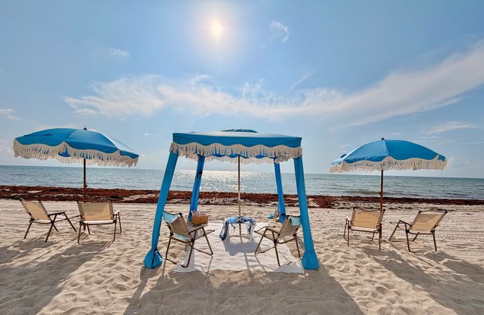 Sunny white-sand beach with fringed blue umbrellas and a central blue canopy over lounge chairs, calm sparkling ocean and bright sunlit sky.