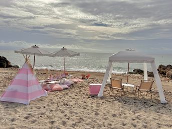 Sunlit beach picnic on a sandy shore with a pink-and-white teepee, low table and cushions, fringe umbrellas, white canopy with folding chairs and a pink cooler facing the sparkling ocean under a cloudy sky.