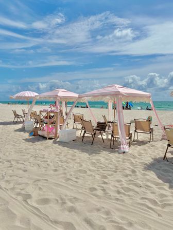 Soft-sand beach with pink-and-white striped cabanas and umbrellas, folding chairs and coolers beside a turquoise ocean under a bright blue sky with wispy clouds — a relaxed summer seaside scene.
