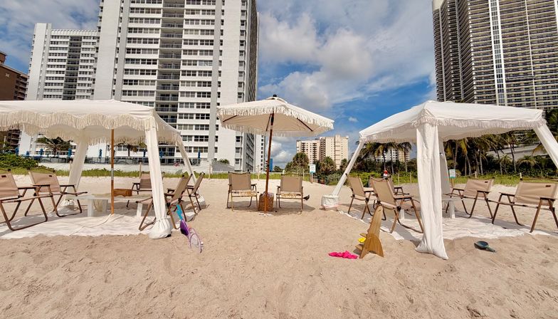 Sunny beachfront setup with fringed white cabanas and umbrella, folding chairs on sand in front of tall oceanfront condos and palm trees under a blue sky